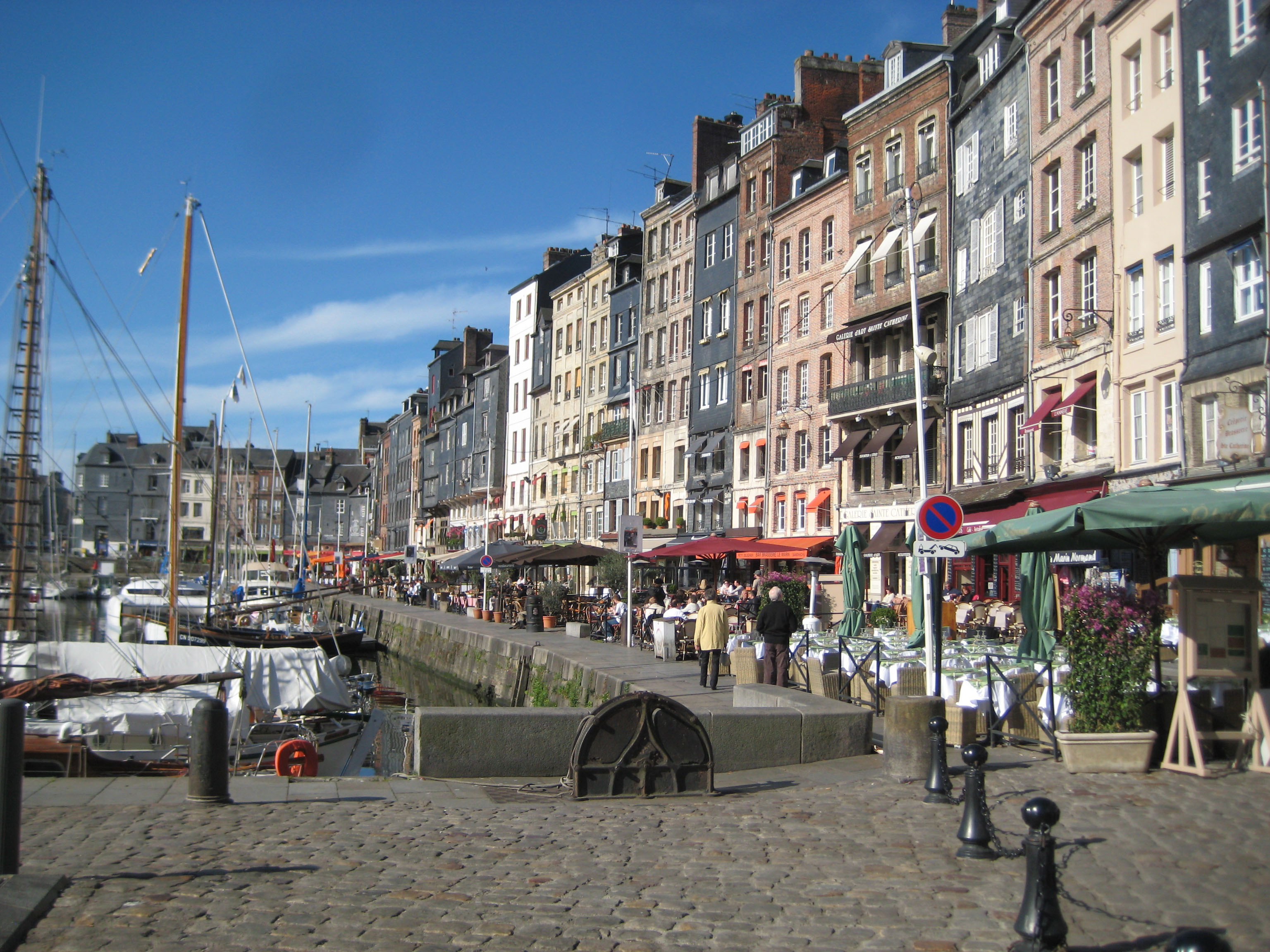 Hamn i Honfleur, Normandie med f�rgglada historiska hus, uteserveringar och b�tar l�ngs kajen under klarbl� himmel.