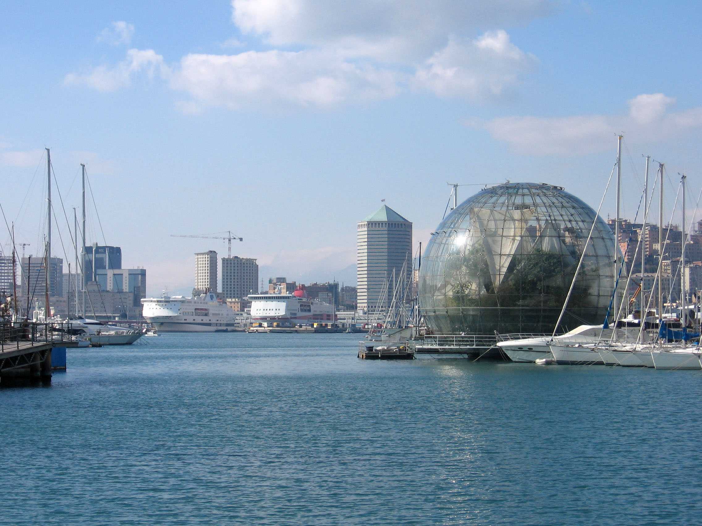 Modern hamn i Genua, Italien med segelb�tar, futuristisk glaskupol och stadens skyline under bl� himmel.
