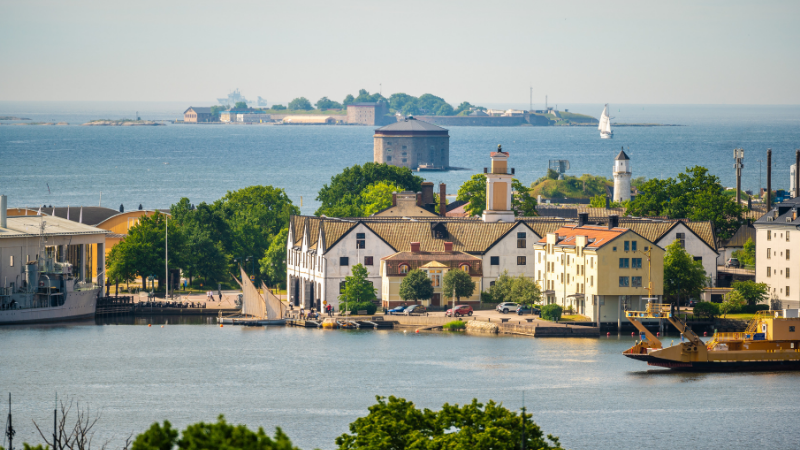 Utsikt över Karlskronas historiska varvsområde med vackra byggnader vid vattnet, skärgårdsöar i bakgrunden och båtar som trafikerar hamnen en solig sommardag.