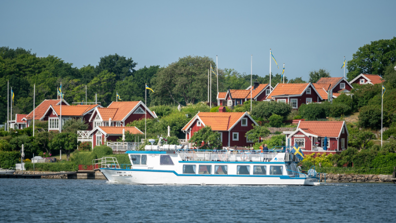 Passagerarbåt i Karlskronas skärgård vid Brändaholm, med röda stugor med vita knutar och svenska flaggor i grönskande sommarlandskap.