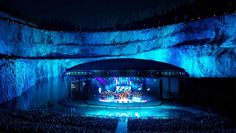 Dalhalla i kv�llsljus med upplyst scen, omgiven av dramatiska kalkstensv�ggar och spegelblank sj�