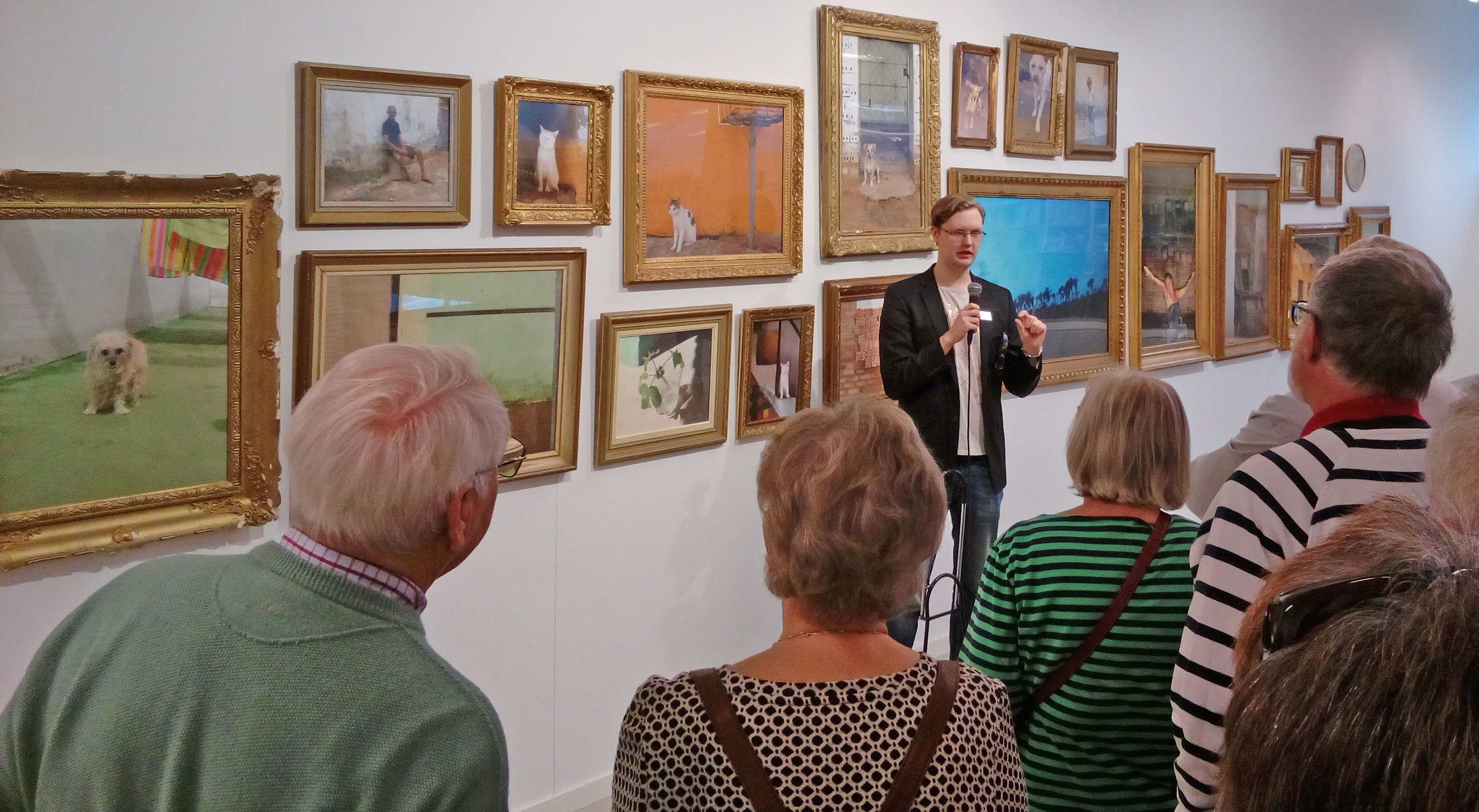 Guidad visning p� Lars Lerins Sandgrund i Karlstad. En grupp bes�kare st�r framf�r en v�gg med flera inramade m�lningar i olika storlekar, f�rest�llande landskap, djur och m�nniskor.