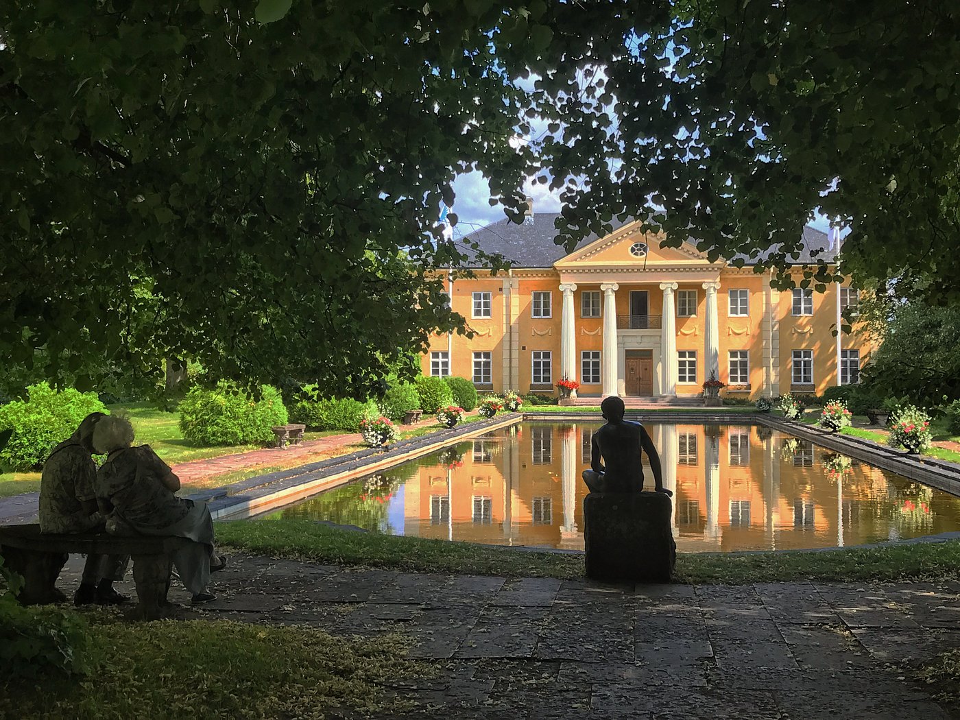 Rottneros Park i Sunne med pampig gul herrgårdsbyggnad i klassisk stil, spegelblank damm och grönskande trädgård. Statyer i förgrunden ramar in den idylliska parkmiljön.