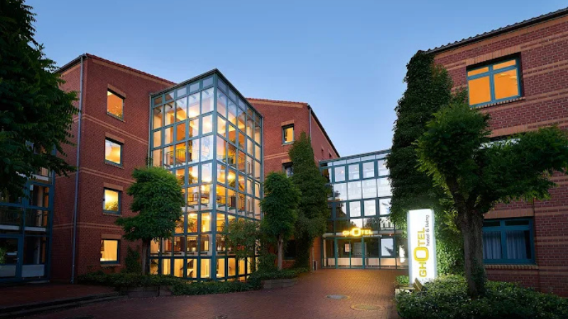 Entré till GHOTEL hotel & living Hannover med modern glasfasad, tegelbyggnader och upplyst skylt i en grönskande innergård.